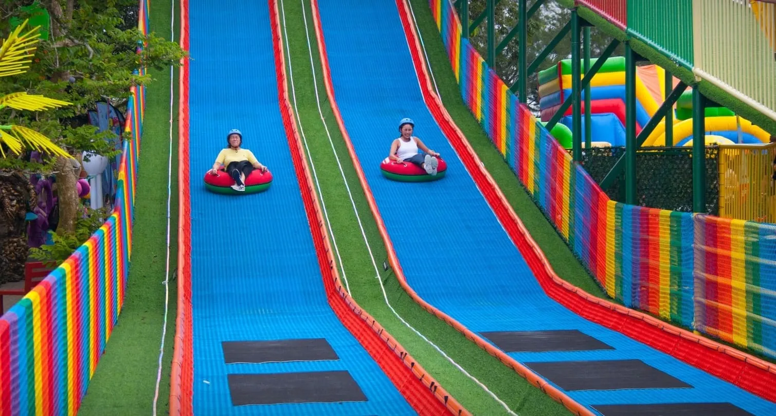 图片展示菲律宾塔格泰 Sky Ranch 内的 Neveplast TUBBY 双滑道，两位游客坐在充气圈中从高处滑下。彩色护栏和蓝色滑道营造出强烈的视觉吸引力，是全年运营的明星项目。