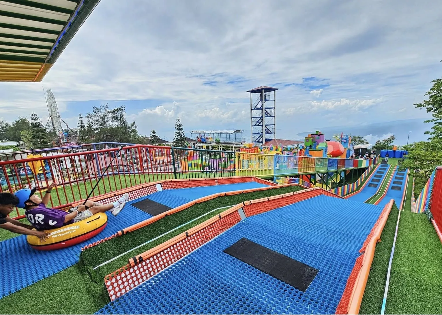 图片展示了 Sky Ranch 内一段设有弯道与缓冲区的 Neveplast TUBBY 滑道，一名游客在工作人员协助下从起点出发。蓝色滑道与绿色草地形成鲜明对比，体现出项目在有限空间内打造丰富滑行线路的灵活性。