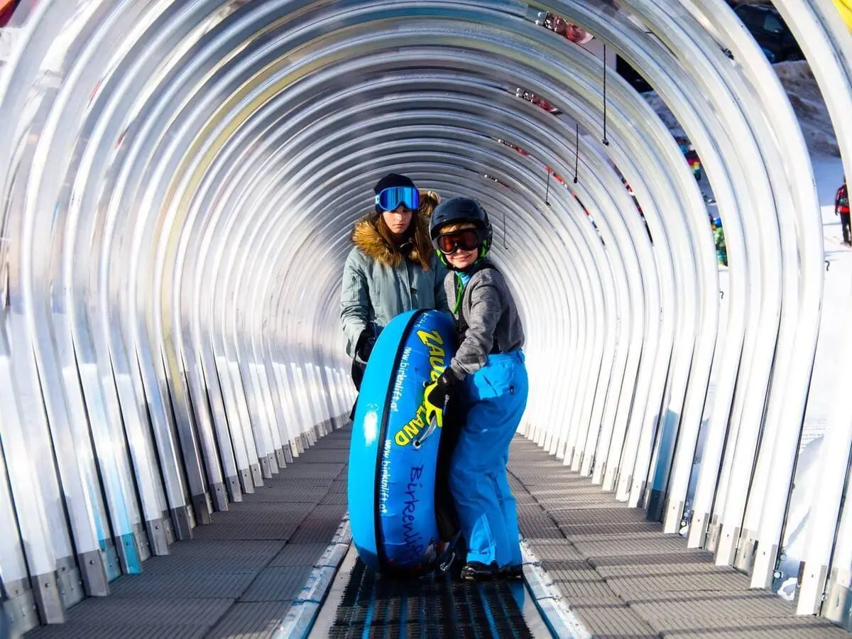 图片中一名儿童坐在蓝色 SNOW TUBES 雪圈上，背景为传送带与滑雪场景，非常适合初学者与儿童活动区域。